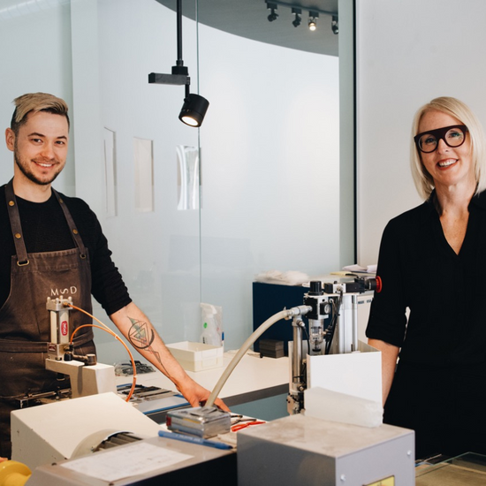 Un artisan lunetier et Marie Sophie Dion souriants dans l’atelier de lunettes à Saint-Lambert, debout près de machines spécialisées pour la fabrication de montures.